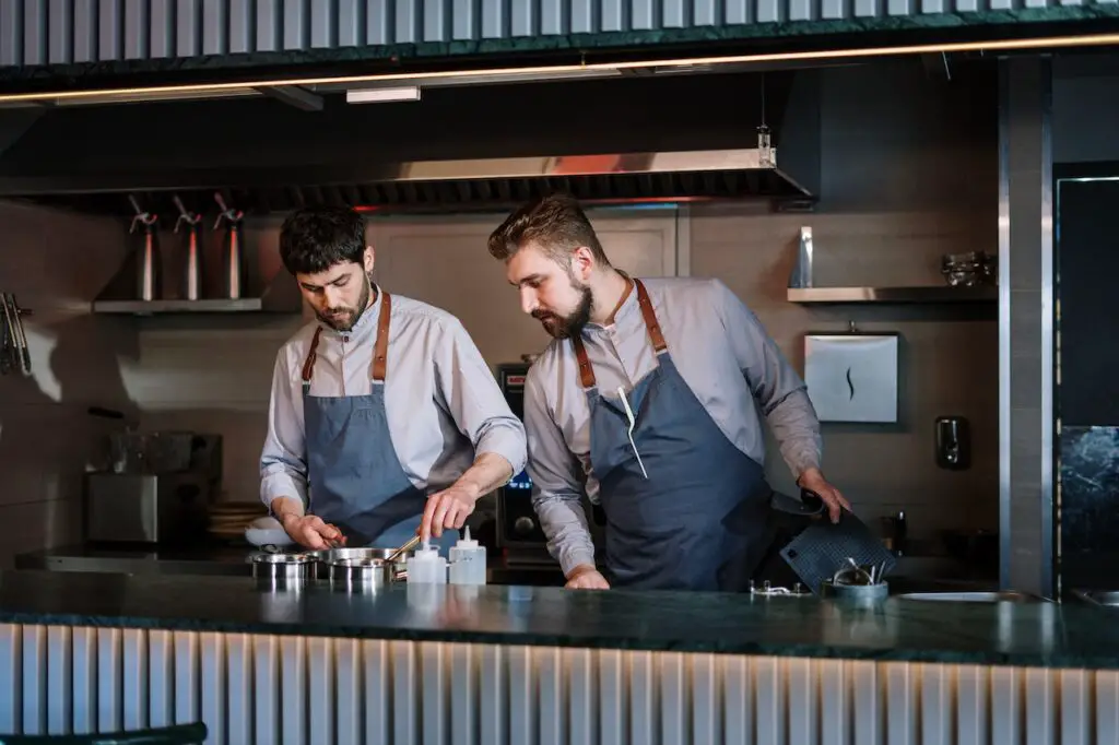 Two men in gray long sleeves and blue aprons cooking on stainless steel in the kitchen