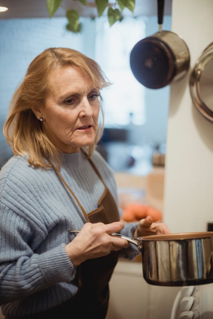 A woman in a blue sweatshirt and a brown apron holds a silver stainless pot in the kitchen