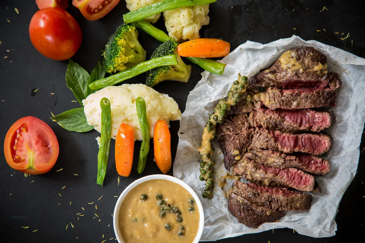 A medium rare steak is placed on parchment paper beside vegetables and sauce in a white bowl