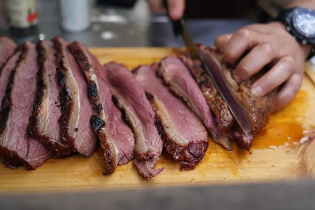 A person wearing a black watch is slicing cooked meat on top of a brown chopping board