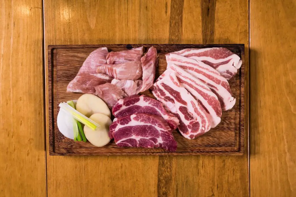Three types of raw meat near a chopped fresh spring onion and peeled potato placed on a wooden chopping board on top of a brown wooden surface