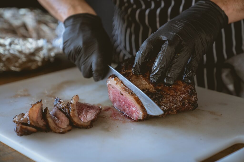 A person wearing black gloves is slicing cooked meat using a silver knife on top of a white chopping board