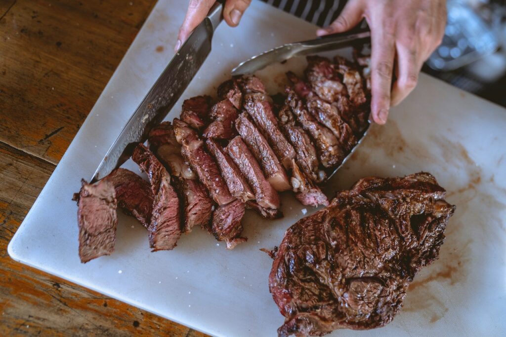 A person is slicing cooked meat using a silver knife and silver tong on a white chopping board