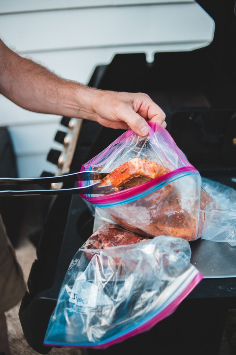 A person putting marinated meat on a resealable plastic bag and silver tongs on top of a black surface