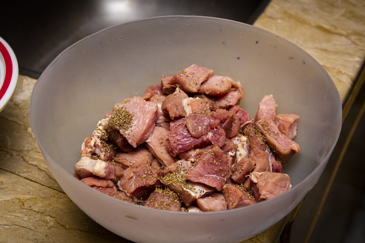 Fresh meat being marinated with herbs in a clear bowl on a marbled countertop