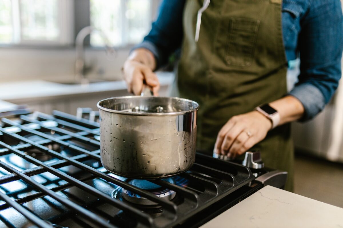 A person wearing a green apron is holding a stainless steel pot placed on top of a black stove