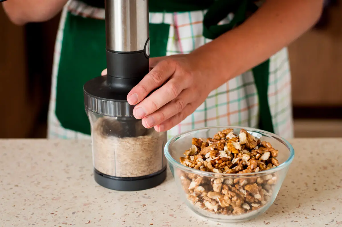 A person wearing a checkered apron is using a black and silver nut chopper near a clear bowl filled with nuts