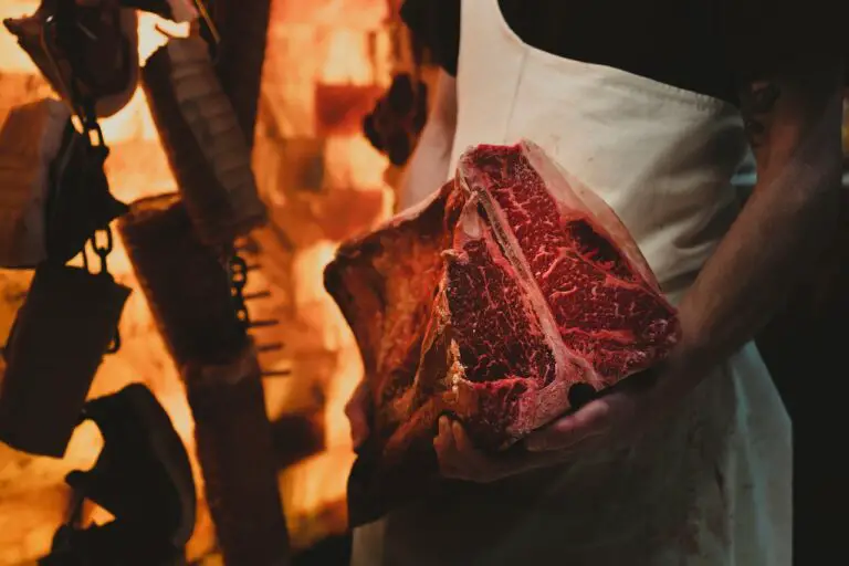 A butcher wearing a white apron and black shirt is holding a slab of dry aged meat