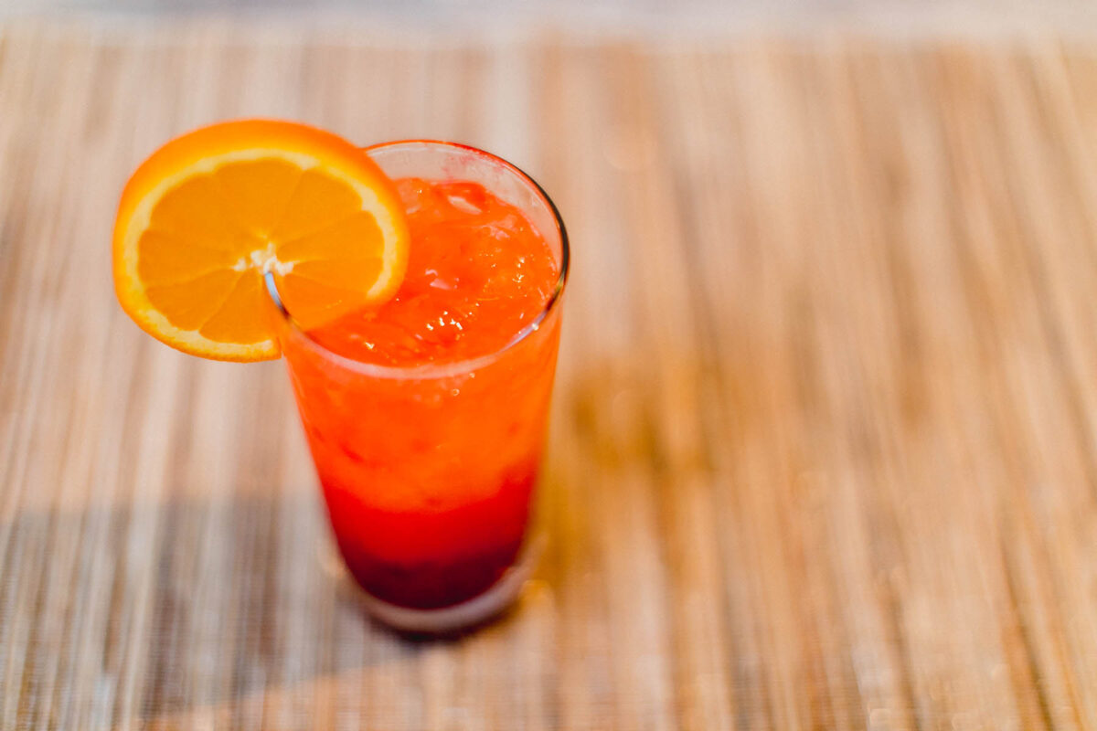 Cranberry tequila sunrise cocktail garnished with an orange slice on a brown placemat