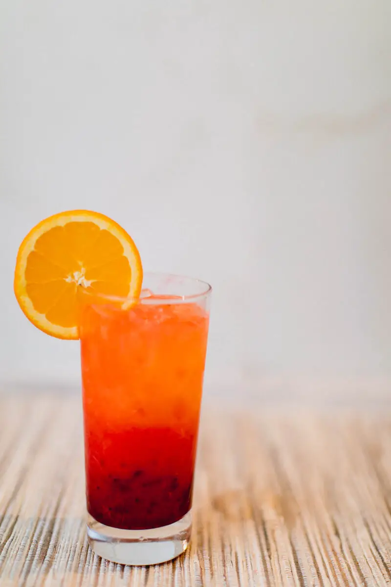 Cranberry tequila sunrise cocktail garnished with a circular orange slide on the side of the glass placed on a light brown surface