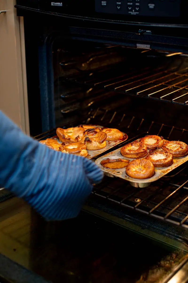 Person using a blue oven mitt to pull 12 popovers out of the oven that are in 2 muffin tins