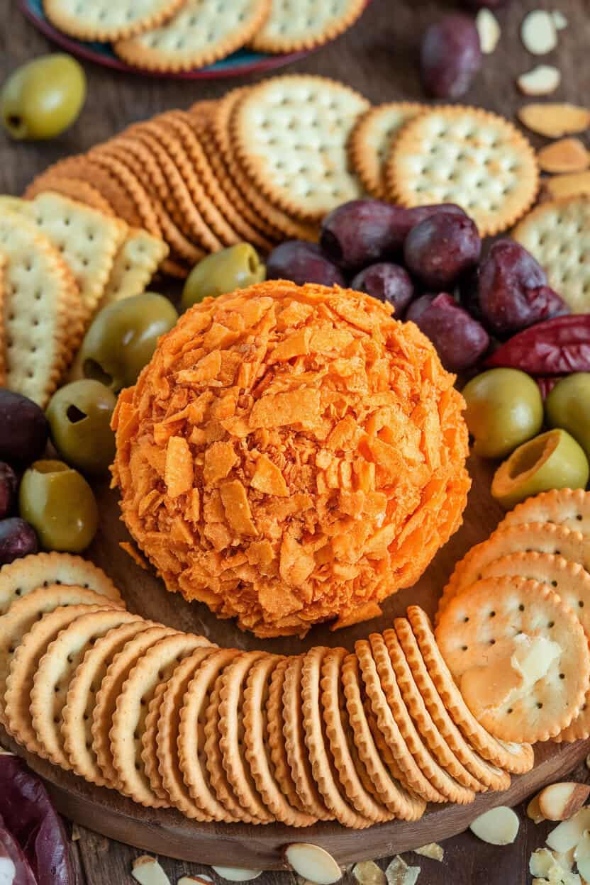 An indoor appetizer board featuring a large orange-tinted cheese ball rolled in crushed nacho chips, surrounded by crackers. No logos or text; photo only.