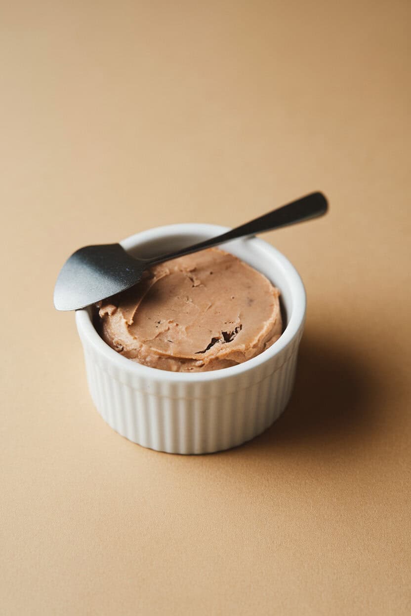 Indoor still life of a small ramekin filled with smooth truffle pâté, tiny spreader resting on the rim, subtle sheen on top. No text or logos.