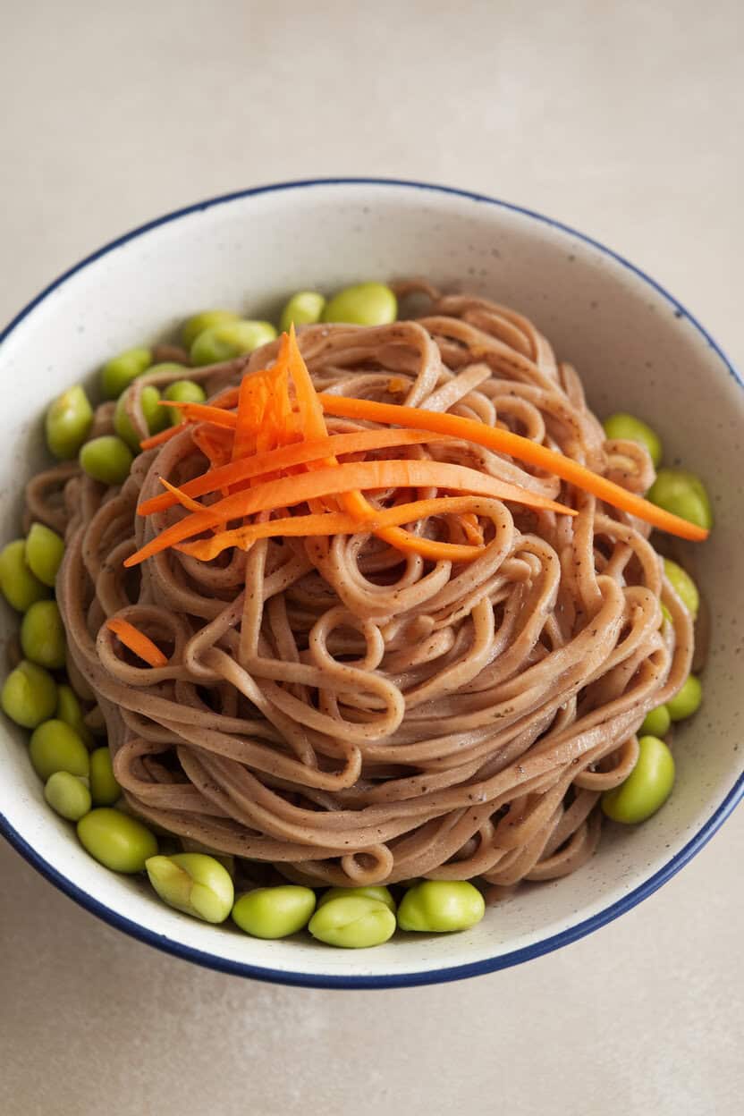 Photo of an indoor bowl brimming with buckwheat soba noodles tossed in a light ginger-garlic sauce, studded with shelled edamame and carrot ribbons. No text or logos.