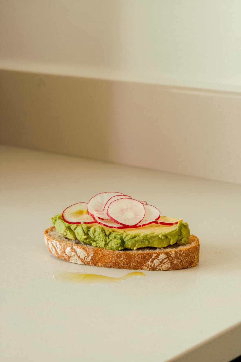 A slice of crusty bread on an indoor countertop topped with smashed avocado, paper-thin radish slices, and a drizzle of olive oil. No text or logos on plate or background.