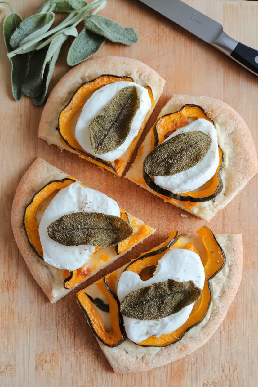 Indoor cutting board holding sliced flatbread topped with roasted acorn squash ribbons, melted mozzarella, and fried sage leaves—overhead shot, no text or logos.