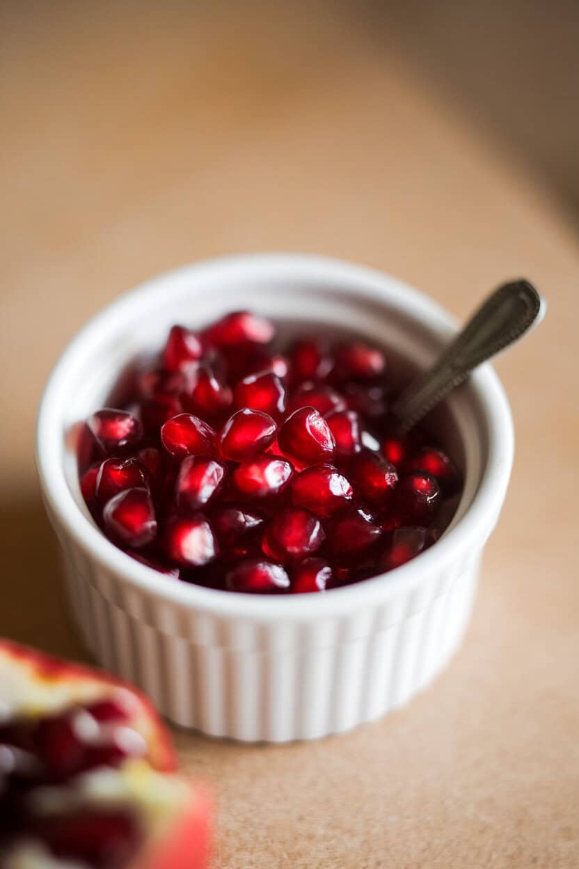 Indoor macro shot of a small ramekin filled with glistening ruby pomegranate seeds, tiny spoon resting alongside. No text or logos.