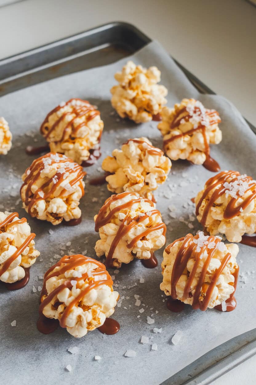 Indoor countertop scene with round popcorn balls drizzled in caramel and sprinkled with flaky salt, set on parchment paper. No text or logos.