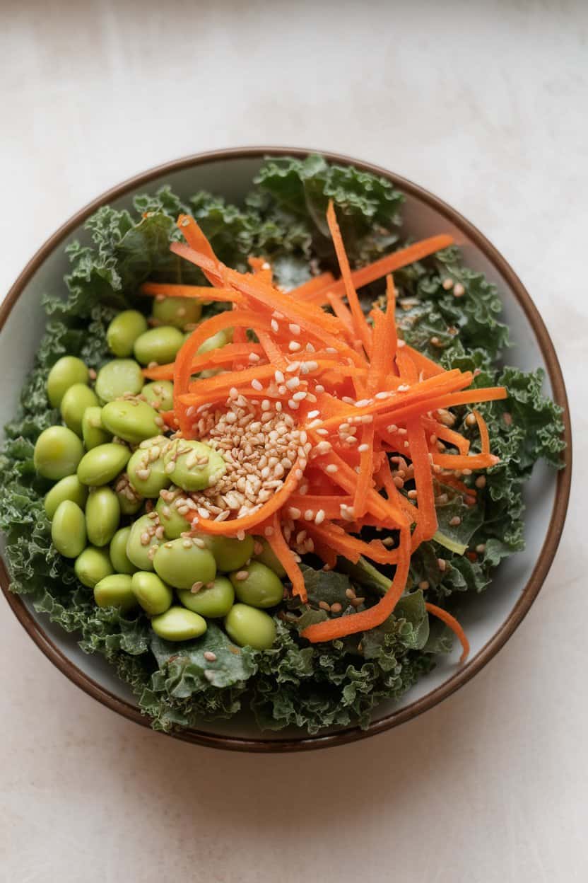 Photo inside a kitchen showing a bowl of massaged kale topped with shredded carrots, edamame, and sesame seeds, lightly dressed with a glossy sesame-ginger vinaigrette. No text or logos visible.