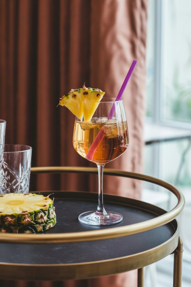 Indoor bar cart displaying a stemmed wine glass of golden spritz with pineapple wedge and purple straw; photo, no text or logos.